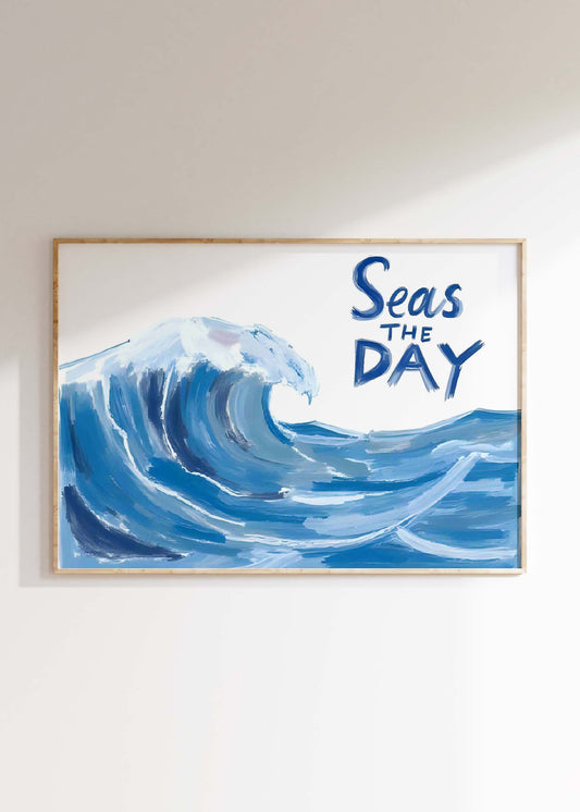 A coastal wall art print in a landscape frame, featuring a large, painterly blue ocean wave and the quote 'Seas the Day'.