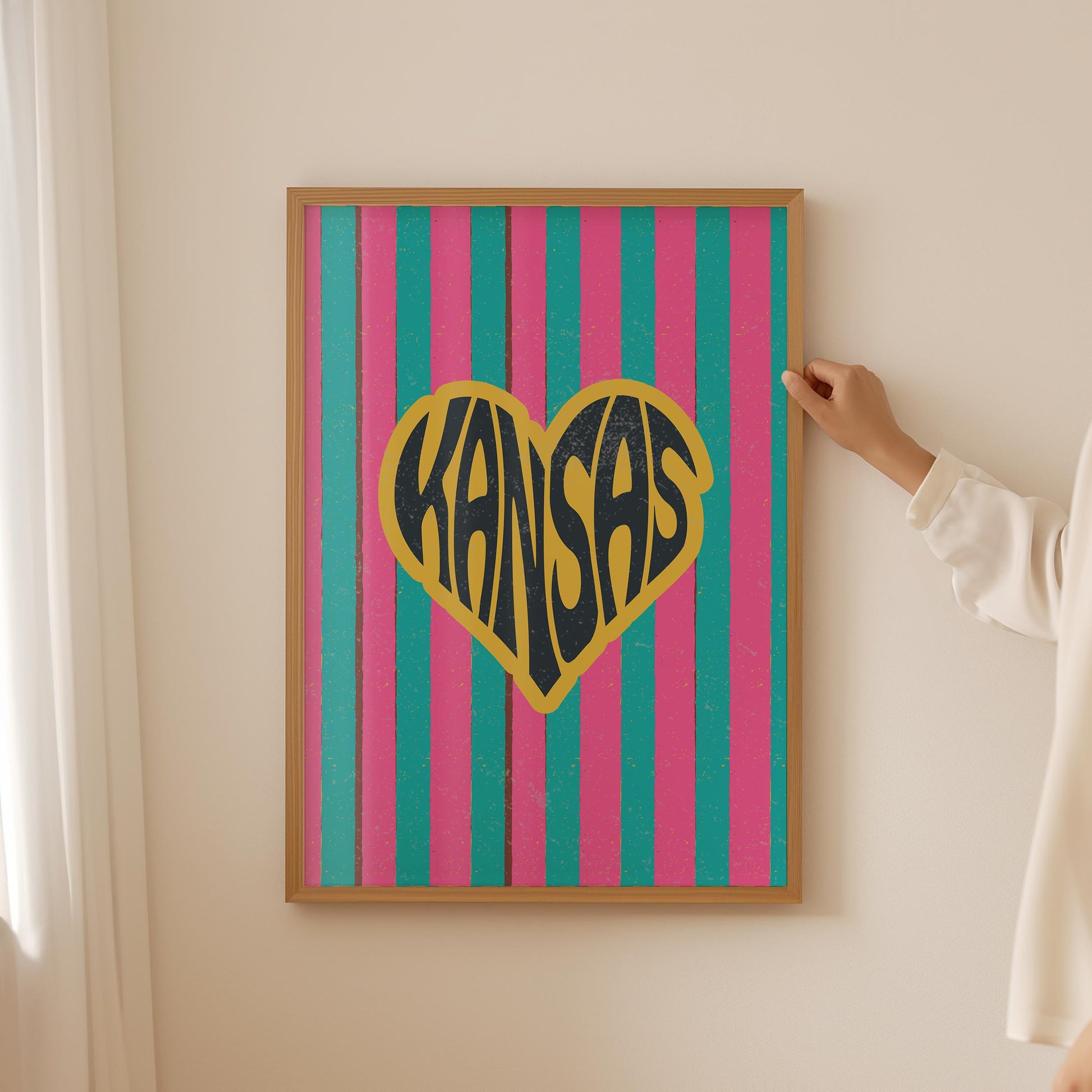 A heart-shaped design with the word 'Kansas' in groovy black retro font, outlined in yellow, set against a background of vertical pink and teal stripes.