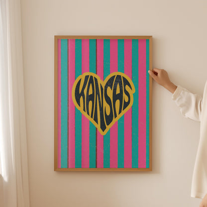 A heart-shaped design with the word 'Kansas' in groovy black retro font, outlined in yellow, set against a background of vertical pink and teal stripes.
