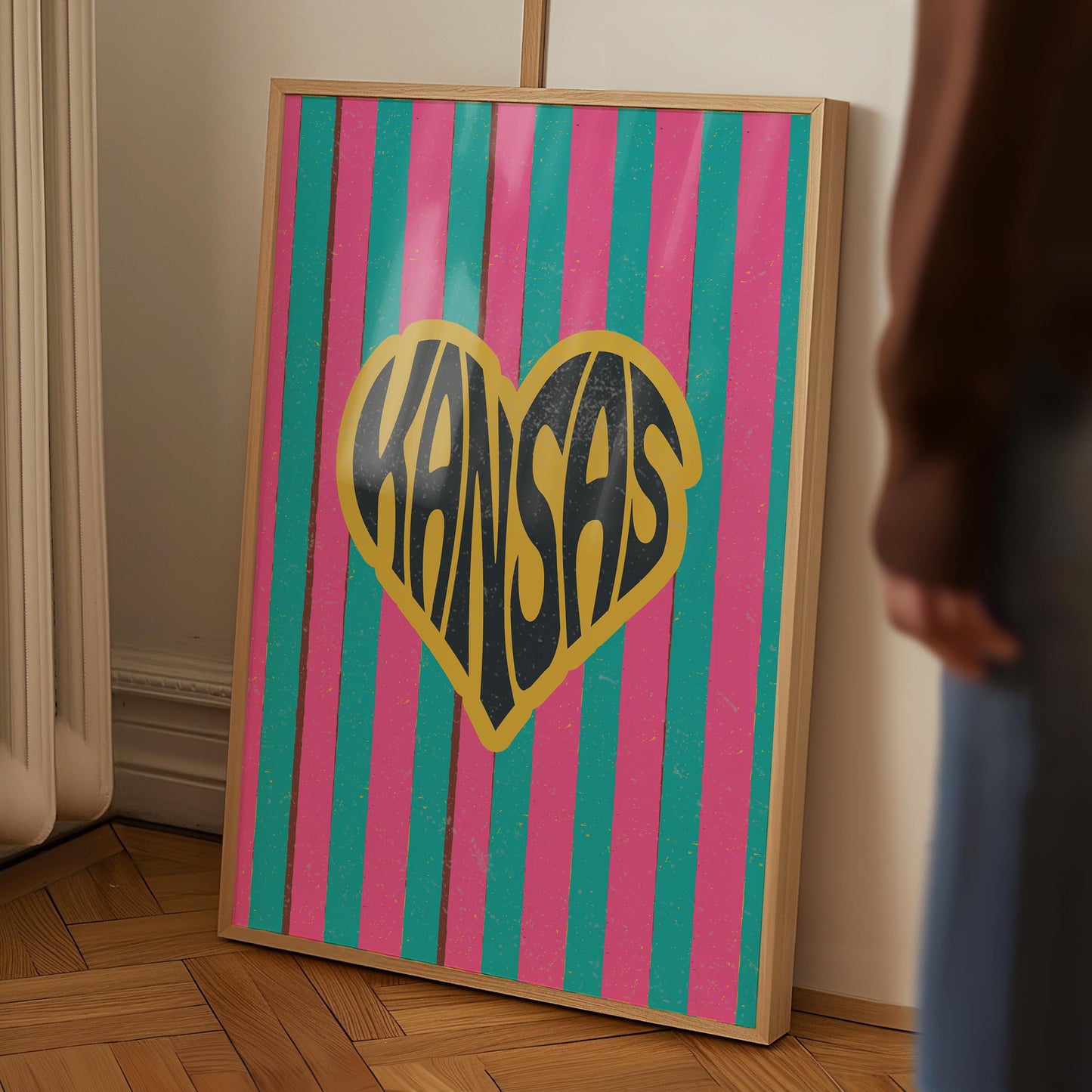 A heart-shaped design with the word 'Kansas' in groovy black retro font, outlined in yellow, set against a background of vertical pink and teal stripes.