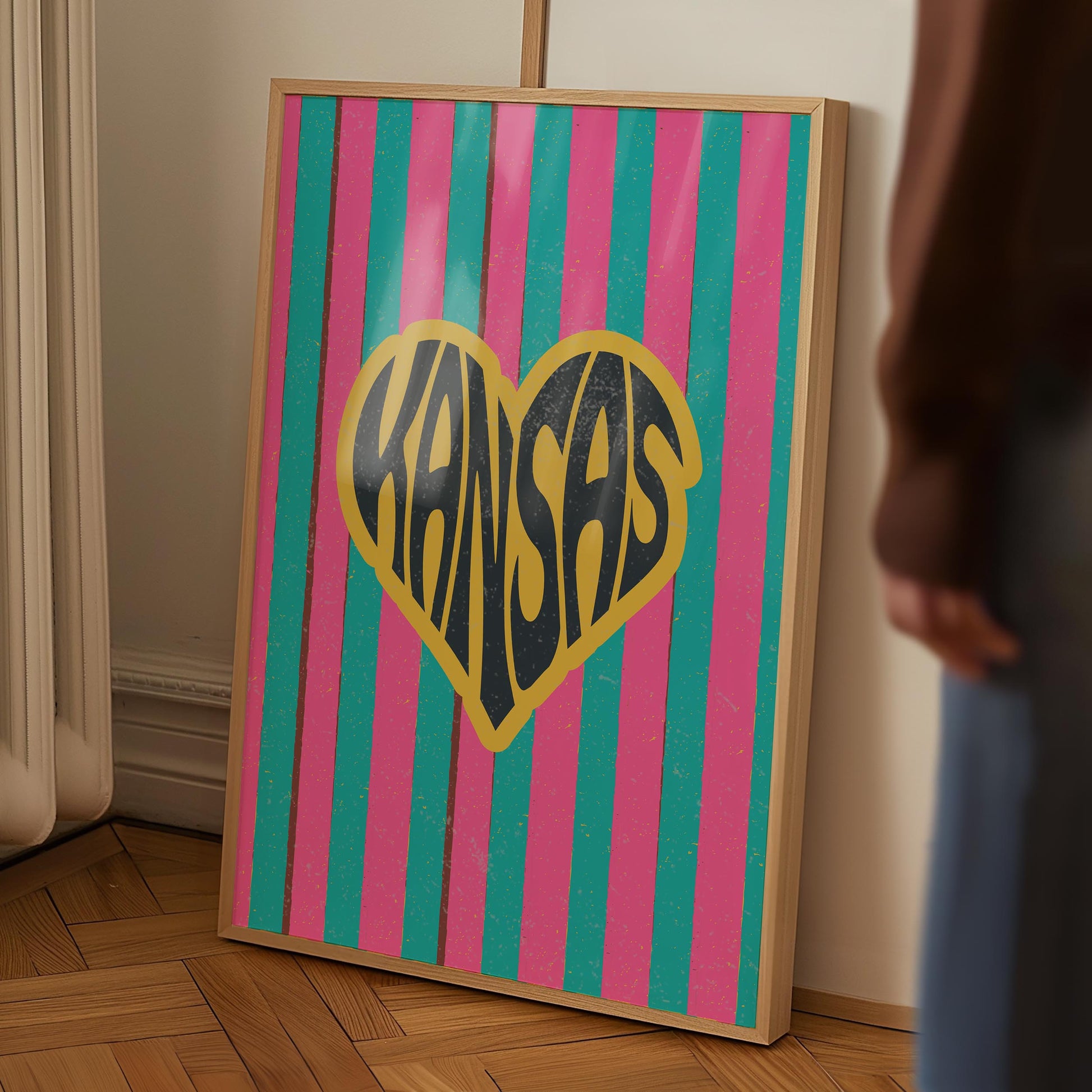 A heart-shaped design with the word 'Kansas' in groovy black retro font, outlined in yellow, set against a background of vertical pink and teal stripes.