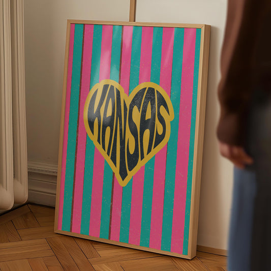 A heart-shaped design with the word 'Kansas' in groovy black retro font, outlined in yellow, set against a background of vertical pink and teal stripes.