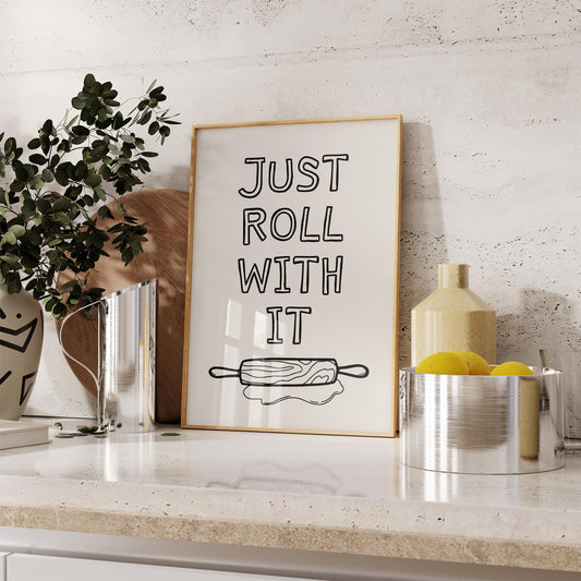 A framed kitchen wall art print with the quote 'Just roll with it' above a hand-drawn illustration of a rolling pin and dough.