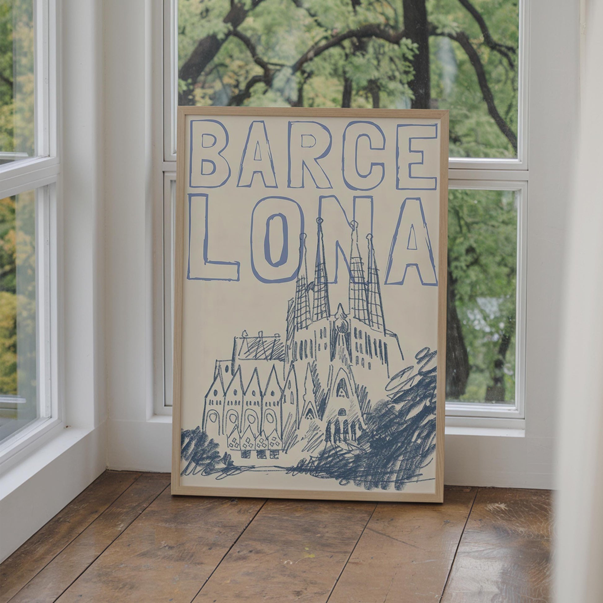A Barcelona travel poster in a wood frame, with a blue sketch of the Sagrada Familia and the word 'BARCELONA' in a block font.